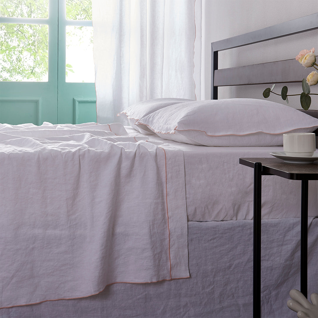 Side angle of 100% linen flat sheet with peach colored edge embroidery laid over a bed