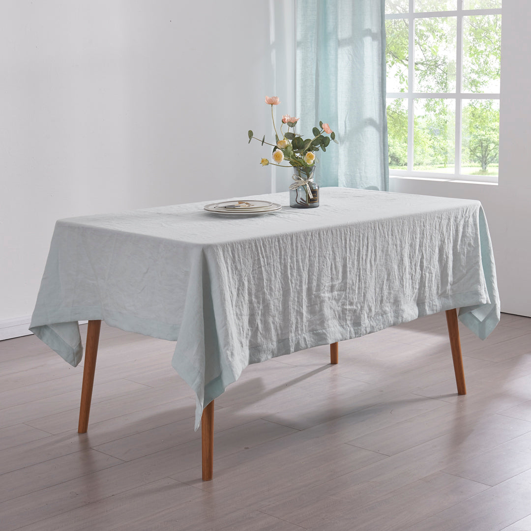 A pale blue 100% linen tablecloth draped over a dining table with a glass flower vase.