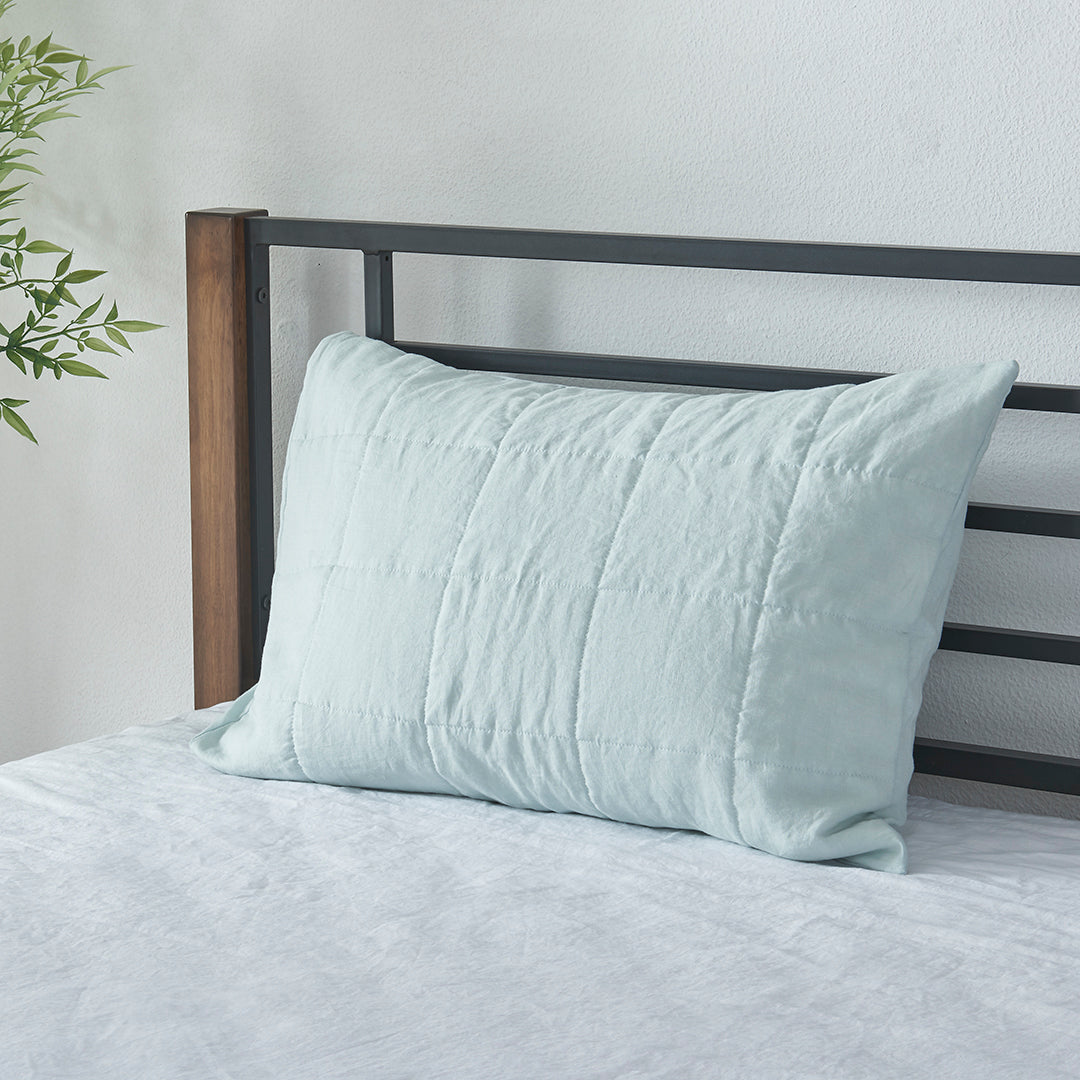 A pale blue 100% linen quilted pillowcase propped up against bed headboard