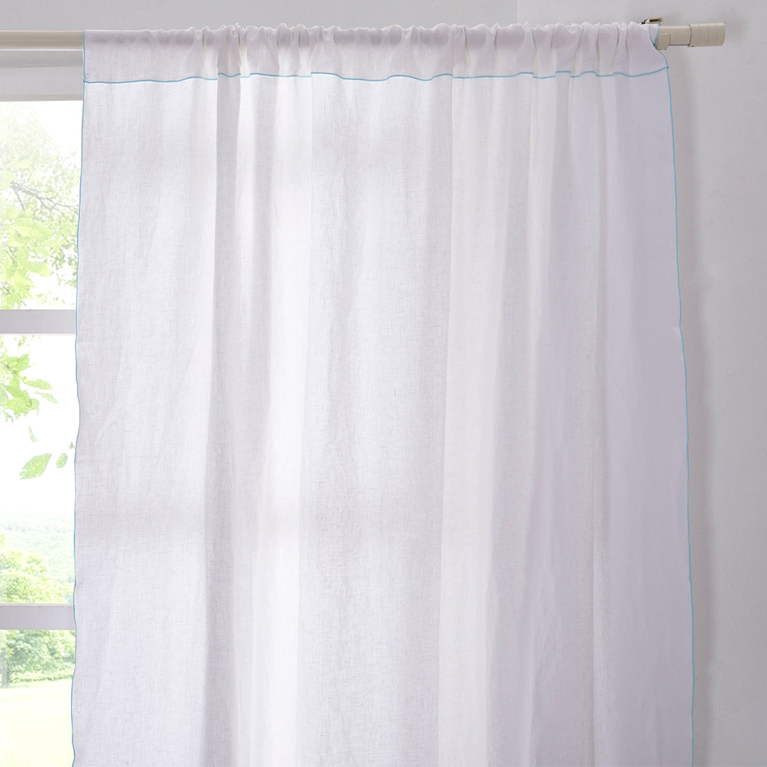 A close-up of a 100% linen curtain with pale blue embroidered edges hanging over a window